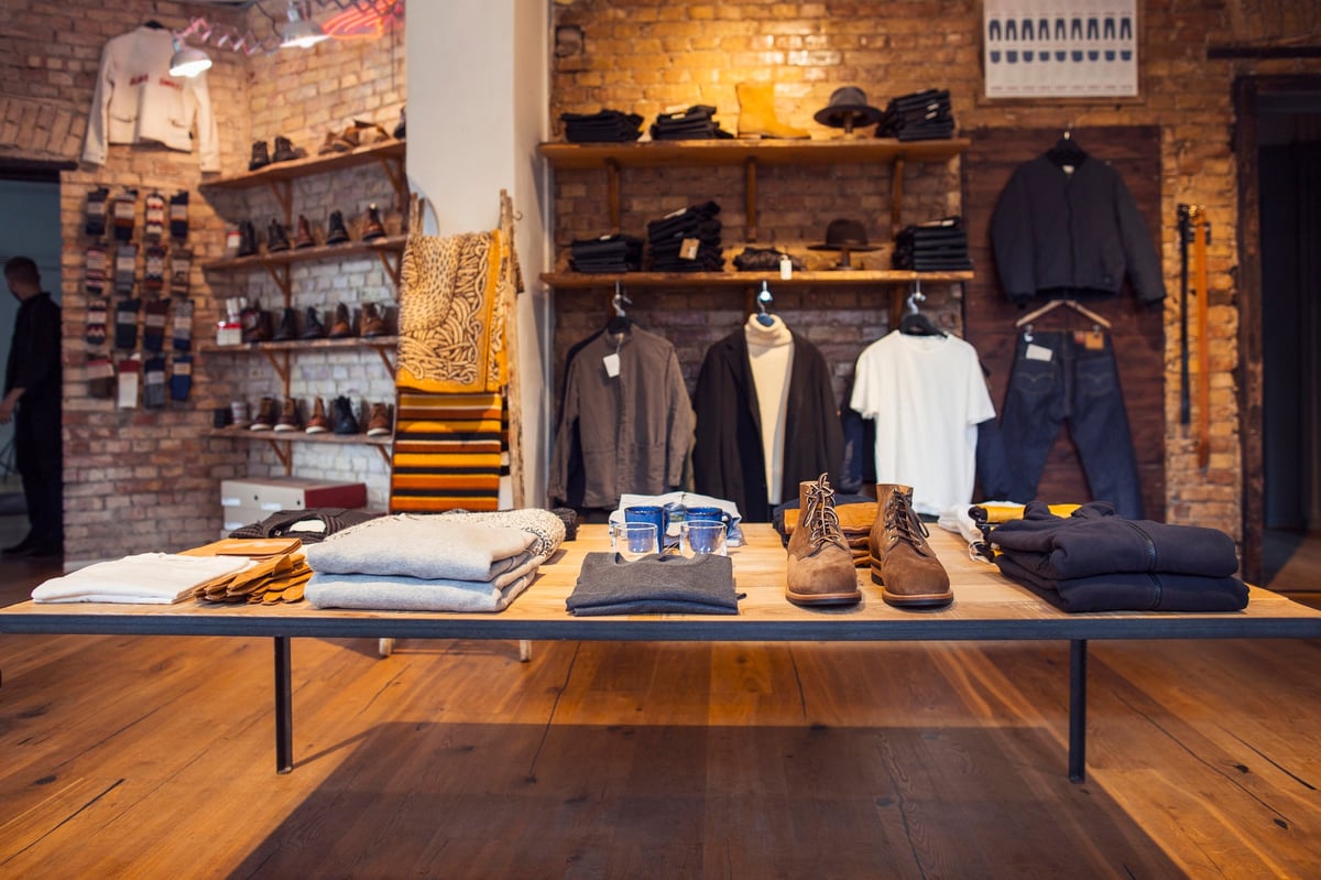 Modern clothing retail display with wooden table featuring folded garments, shoes, and accessories against exposed brick wall with hung clothing and shelving