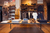 Modern clothing retail display with wooden table featuring folded garments, shoes, and accessories against exposed brick wall with hung clothing and shelving