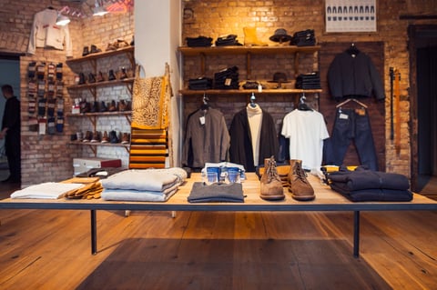 Modern clothing retail display with wooden table featuring folded garments, shoes, and accessories against exposed brick wall with hung clothing and shelving