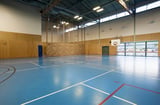 Empty indoor gymnasium with blue court markings, wooden walls, and natural light from windows