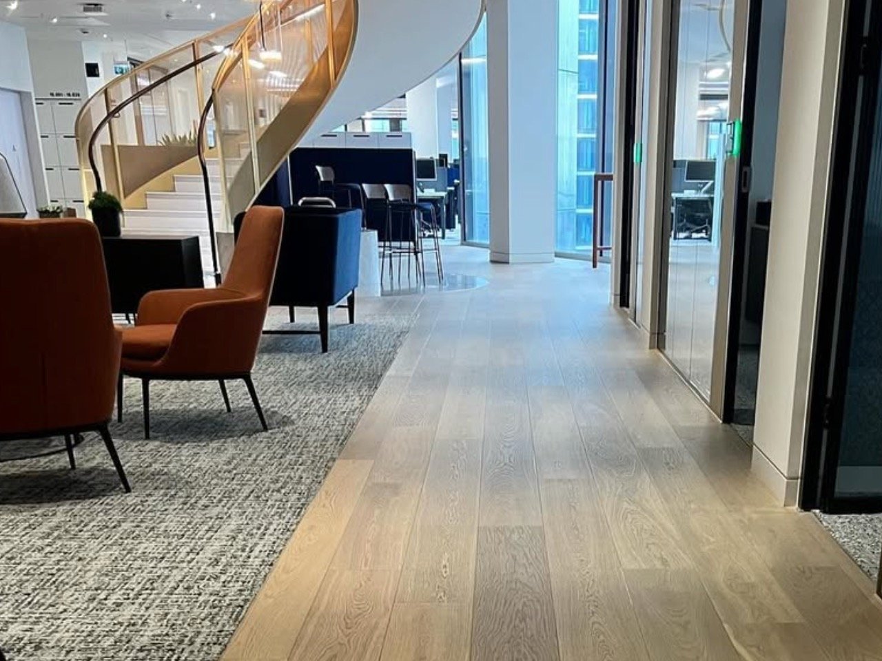 Modern office hallway with light wood flooring, lounge seating, wooden staircase, and floor-to-ceiling windows overlooking city buildings