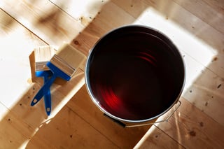Overhead view of an open paint can with dark red paint and a blue paint brush on wooden floor with sunlight