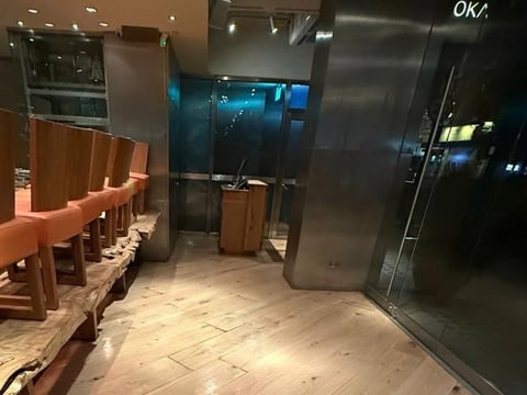 Modern restaurant interior with wooden chairs, beige tile flooring, and dark glass and metal fixtures, dimly lit with blue city lights visible through windows