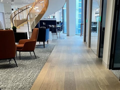 Modern office hallway with wooden flooring, orange chairs, staircase, and floor-to-ceiling windows overlooking city buildings