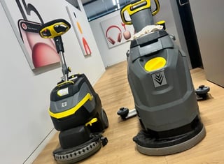 Two industrial floor cleaning machines with yellow and black accents displayed in a showroom with wall-mounted product displays
