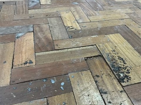 Close-up of damaged hardwood floor with herringbone pattern, showing water stains, dark discoloration, and worn finish.