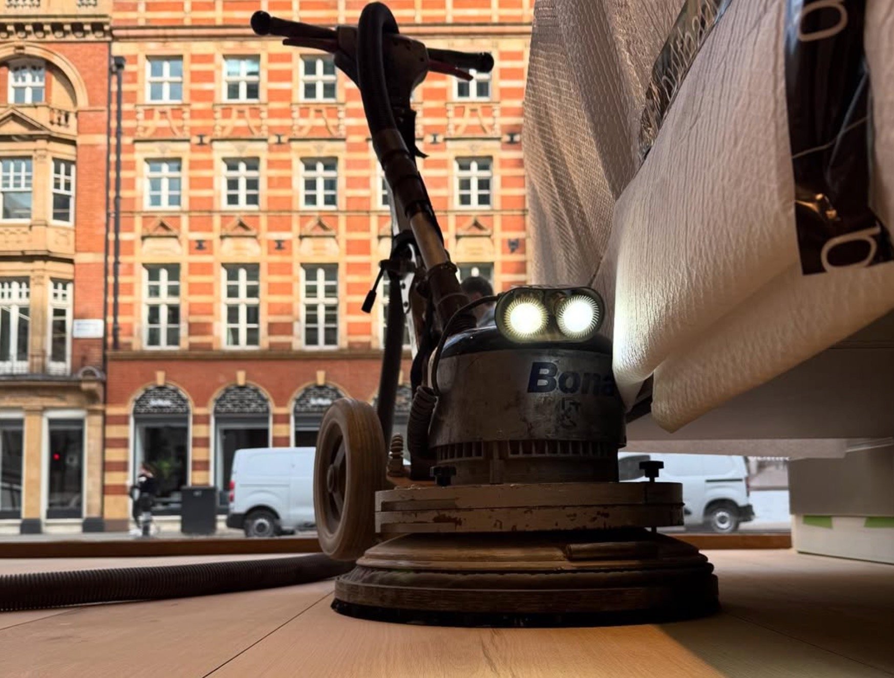 Toy street cleaning machine with illuminated headlights positioned on wooden surface with urban building backdrop